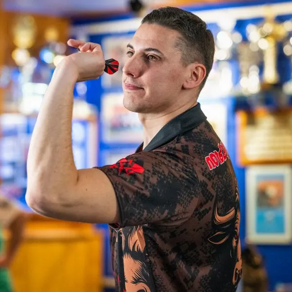 Ein Mann in einem gemusterten Sporttrikot mit roten Akzenten hält einen Dart und zielt, im Hintergrund sind Trophäen zu sehen.