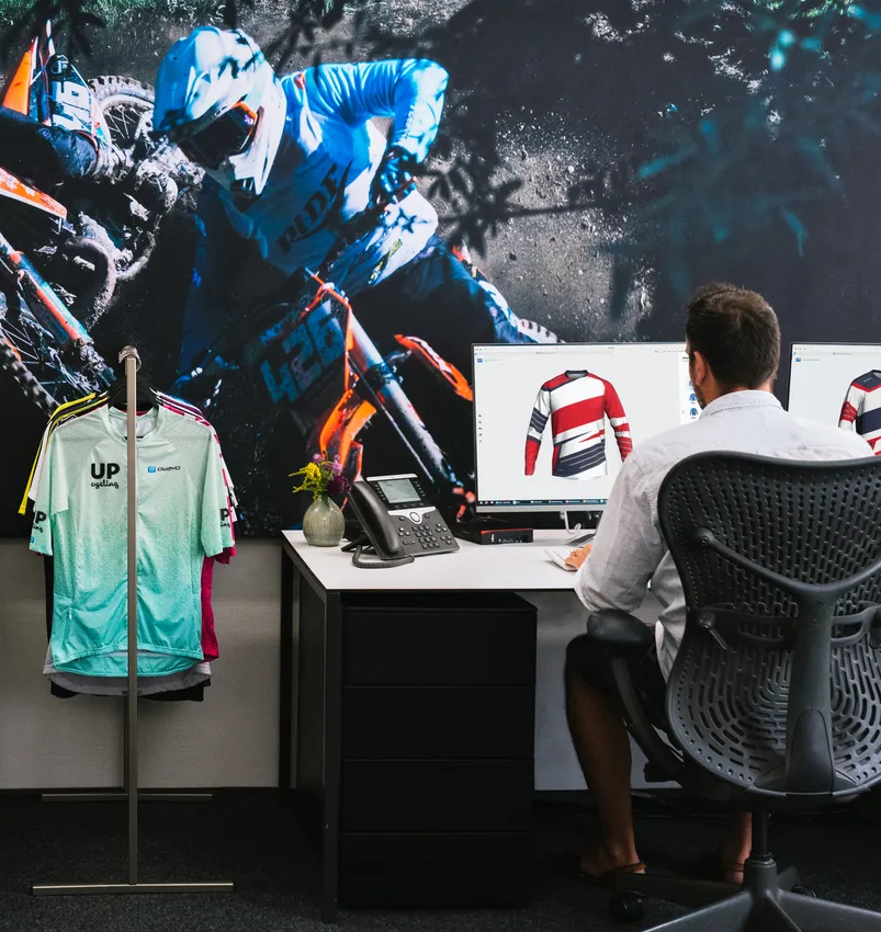 Ein Mann sitzt an einem Schreibtisch und entwirft auf einem Computer ein maßgeschneidertes Sporttrikot, während im Hintergrund ein großes Bild eines Motorradfahrers und ein Kleiderständer mit verschiedenen Sportbekleidungsstücken zu sehen sind.
