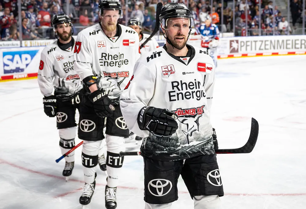 Eishockeyspieler in einem weißen Trikot mit schwarzen Hosen, die das Logo eines Teams tragen, während sie auf dem Eis stehen.