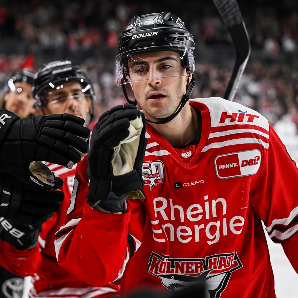 Ein Eishockeyspieler in einem roten Trikot mit weißen Streifen und Logos, der seine Hand zum Feiern hebt.