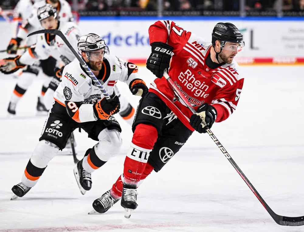 Ein Eishockeyspieler in rotem Trikot und schwarzer Hose fährt auf dem Eis und wird von einem anderen Spieler verfolgt.