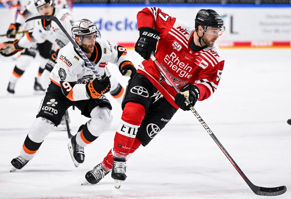 Ein Eishockeyspieler in einem roten Trikot fährt mit dem Puck über das Eis, während ein anderer Spieler in einem weißen Trikot ihm folgt.