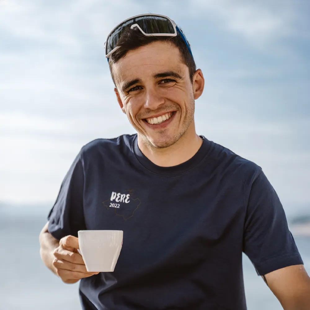 Ein Mann hält eine Tasse in der Hand und trägt ein dunkelblaues T-Shirt mit dem Aufdruck 'PERE 2022', lächelnd vor einem verschwommenen Hintergrund.