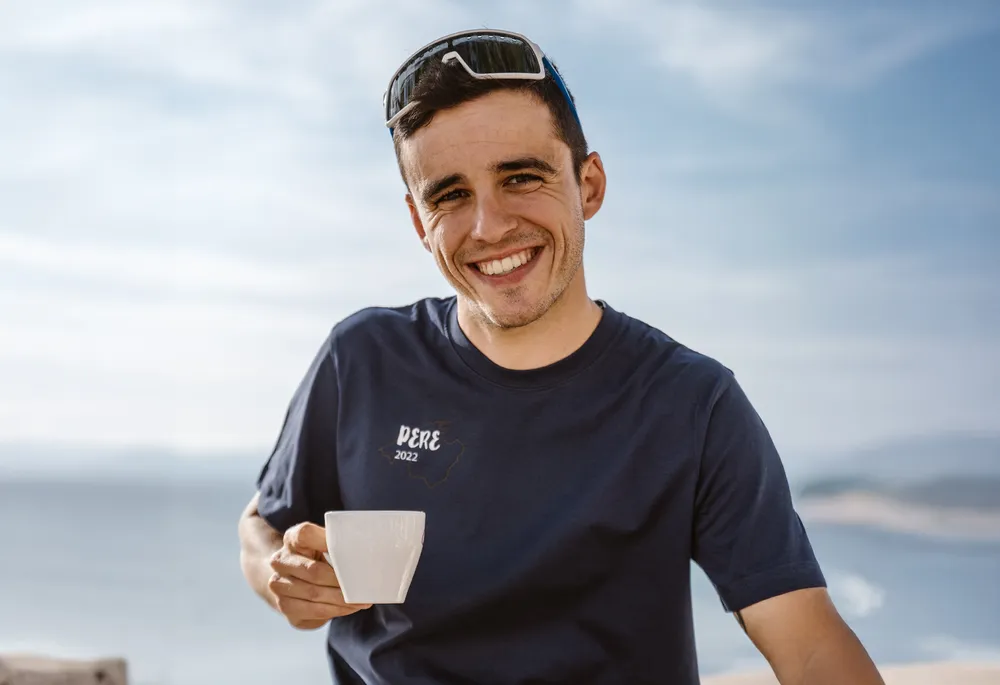 Ein lächelnder Mann in einem blauen T-Shirt hält eine Tasse in der Hand, vor einer schönen Küstenlandschaft.