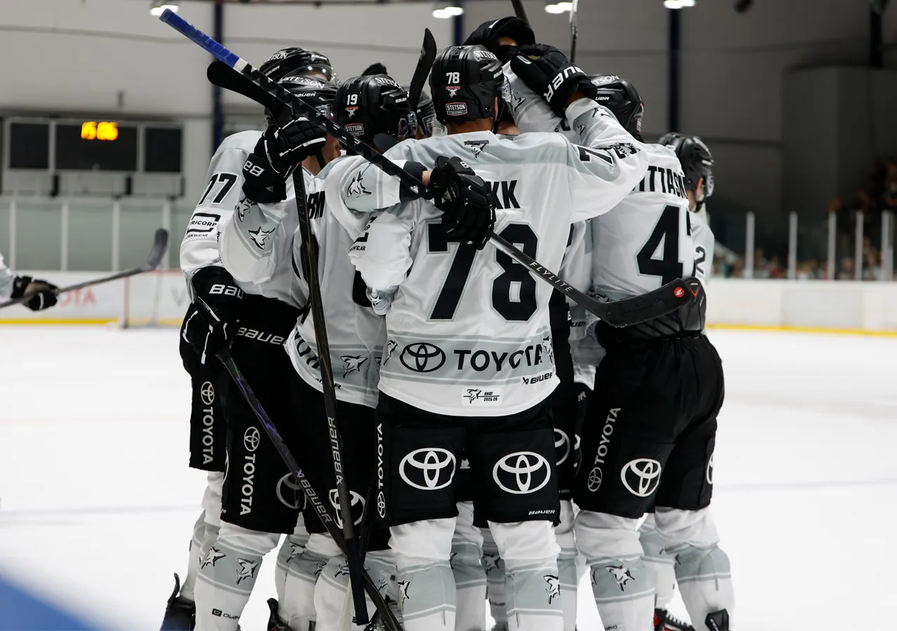Eishockeyspieler in weißen Trikots mit schwarzen Nummern und Sponsorenlogos, die sich auf dem Eis umarmen.