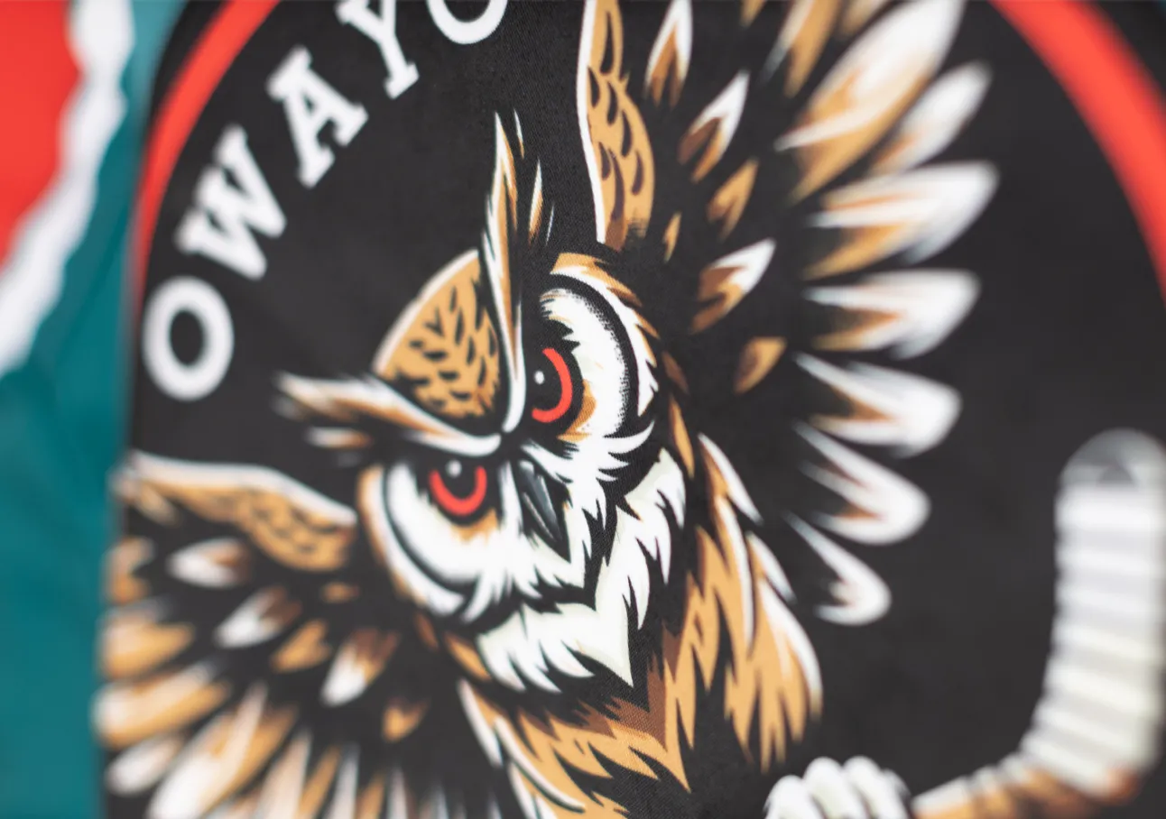 Ein stilisierter Eulenkopf mit roten Augen auf einem schwarzen Hintergrund, umgeben von einem roten und weißen Kreis, auf einem Sportbekleidungsstück von owayo.