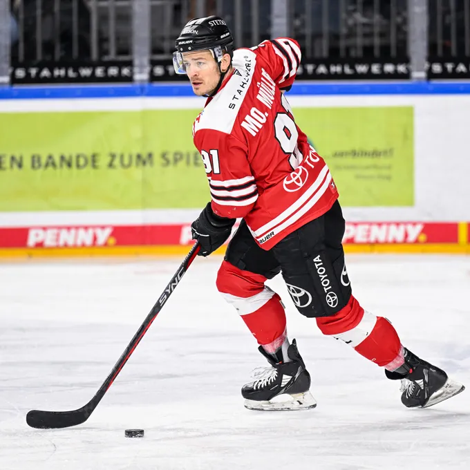 Ein Eishockeyspieler in einer roten und schwarzen Trikot mit der Nummer 91 und dem Namen Mc. Miller auf dem Rücken, der auf dem Eis steht und einen Puck mit seinem Schläger kontrolliert.