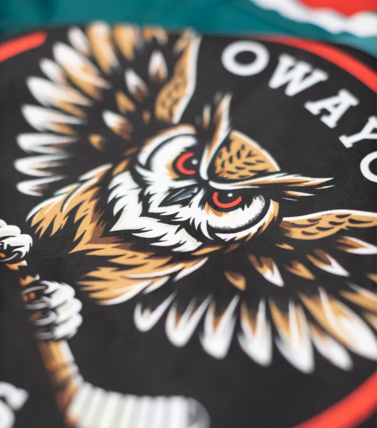 Eine Nahaufnahme eines schwarzen Sporttrikots mit einem stilisierten Eulen-Logo, das einen Eishockeyschläger hält, und dem Schriftzug 'owayo' in weißer Schrift.