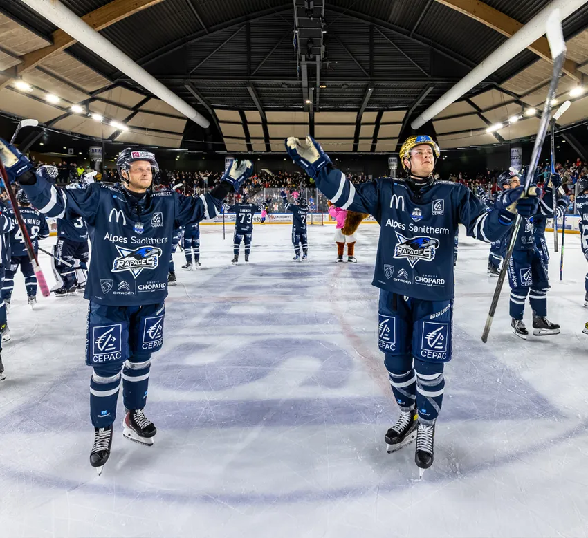 Eishockeyspieler in blauen Trikots mit Sponsorenlogos feiern auf dem Eis, während im Hintergrund das Publikum zu sehen ist.