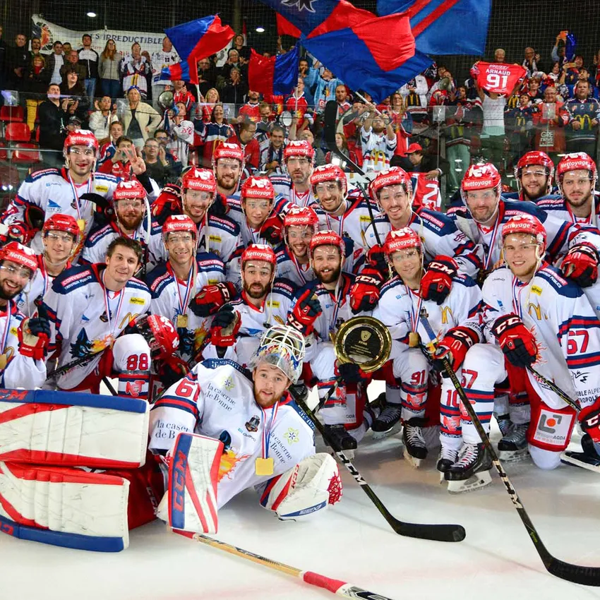 Eine Eishockeymannschaft posiert stolz auf dem Eis mit Medaillen und einem Pokal, während Fans im Hintergrund jubeln.