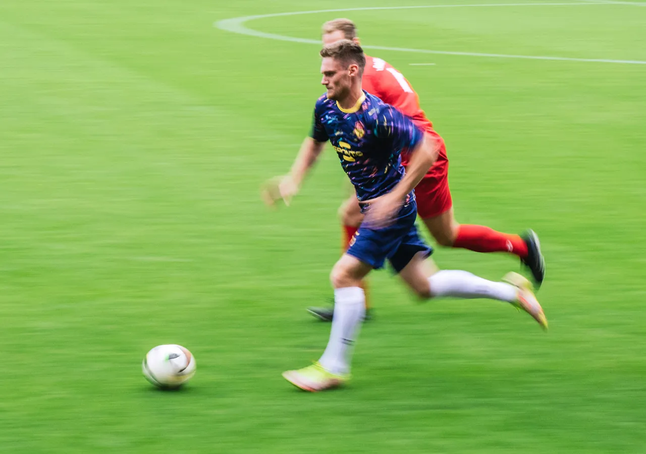 Ein Fußballspieler in einem bunten Trikot sprintet über den Rasen, während ein anderer Spieler ihm nahe folgt.