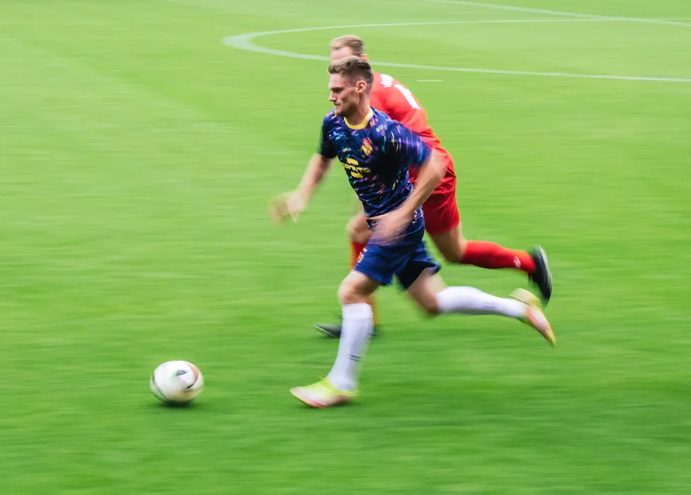 Ein Fußballspieler in einem bunten Trikot sprintet über den Rasen, während ein anderer Spieler ihm folgt.