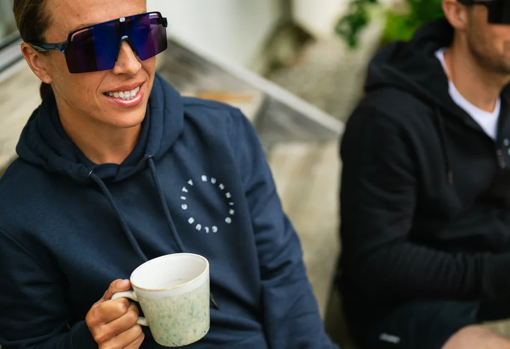 Eine Person in einem blauen Hoodie mit einem Logo hält eine Tasse in der Hand, während sie auf einer Holzbank sitzt.