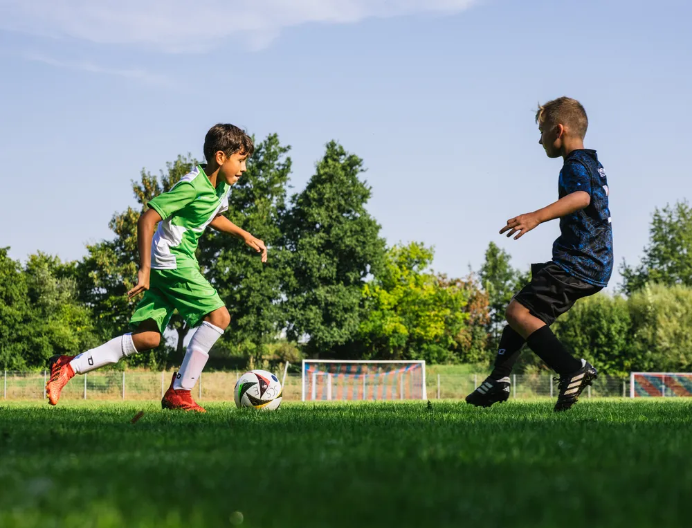Zwei Kinder spielen Fußball auf einem grünen Feld, eines trägt ein grünes Trikot und das andere ein schwarzes, während sie den Ball verfolgen.