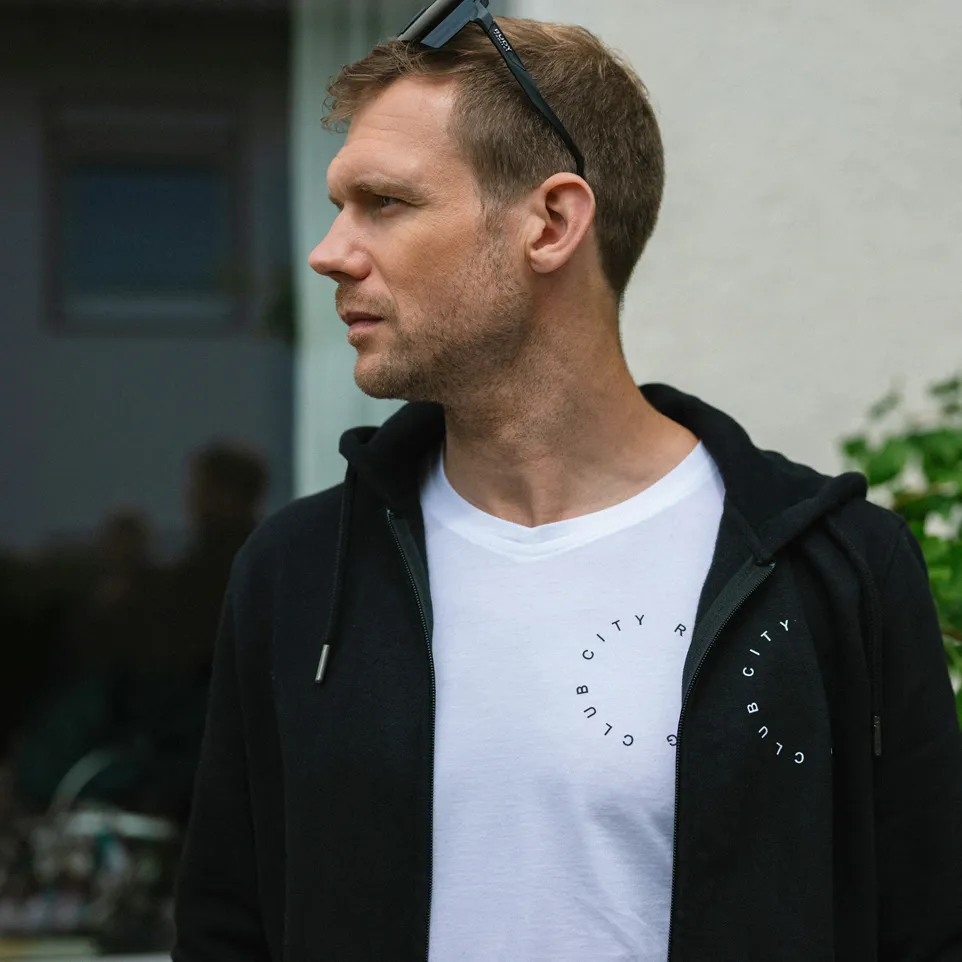 Ein Mann trägt ein schwarzes Hoodie über einem weißen T-Shirt mit einem runden Aufdruck.