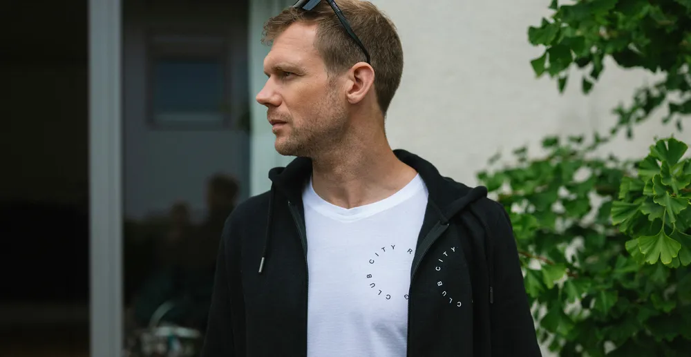 Ein Mann steht seitlich und trägt ein schwarzes Hoodie über einem weißen T-Shirt mit einem Aufdruck, während er von Pflanzen umgeben ist.