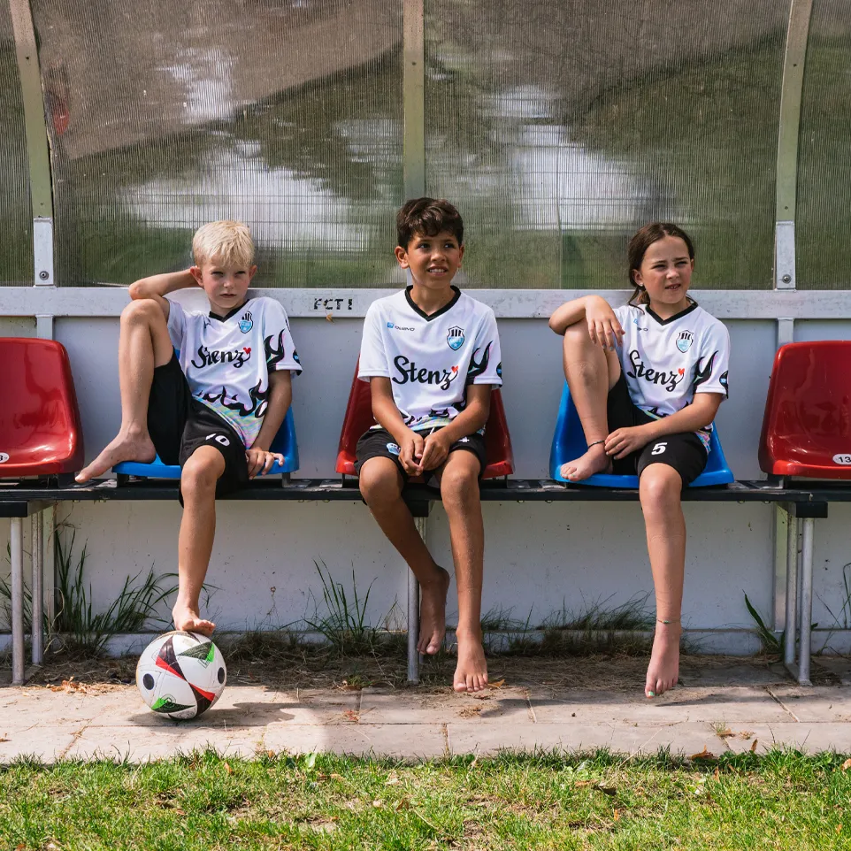 Drei Kinder in Fußballtrikots und Shorts sitzen auf einer Bank am Spielfeldrand, während ein Fußball vor ihnen liegt.