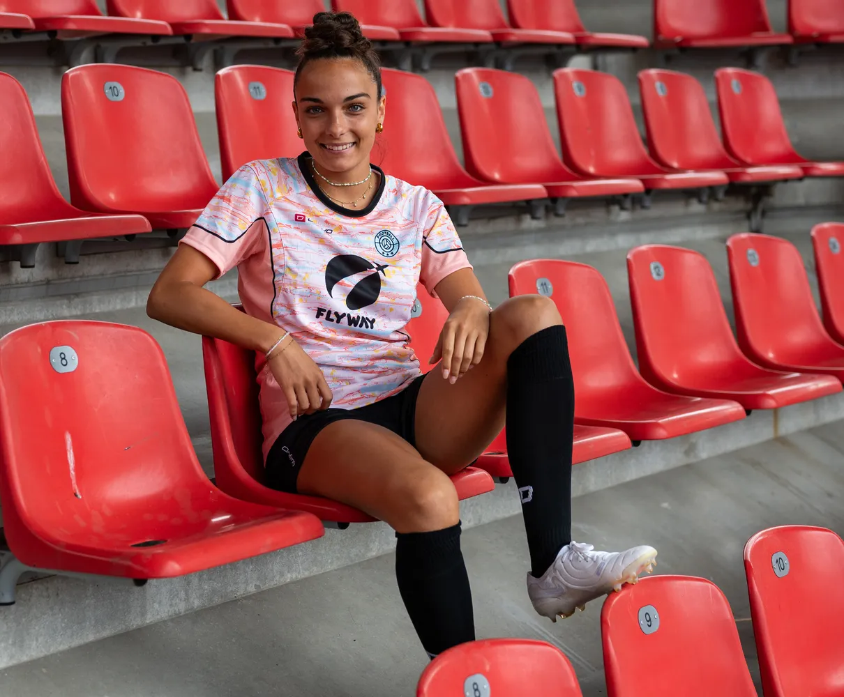 Eine junge Frau sitzt auf einer roten Tribüne in einem bunten Fußballtrikot und schwarzen Socken.