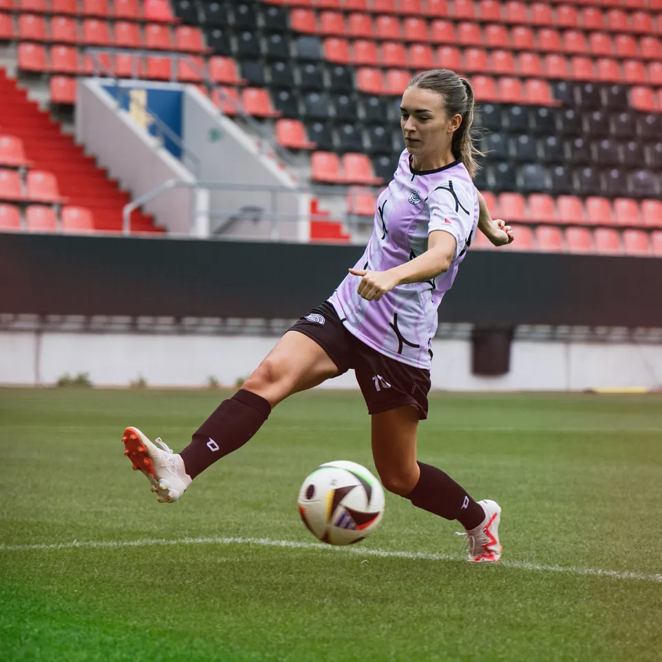 Eine Spielerin tritt einen Fußball auf einem Stadionrasen, bekleidet mit einem lila Trikot und schwarzen Shorts.