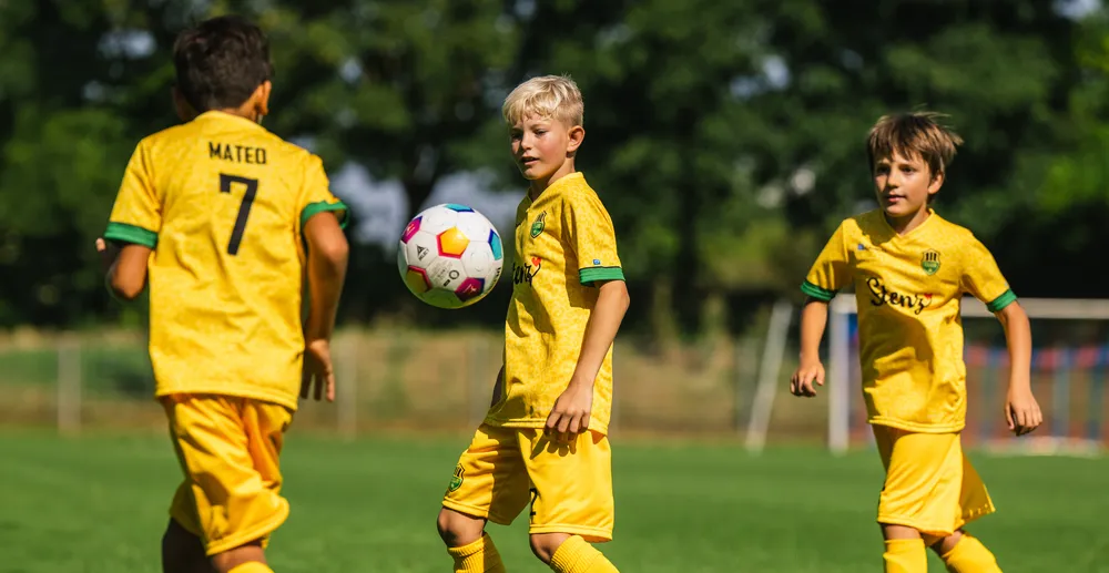 Drei Kinder in gelben Fußballtrikots spielen auf einem grünen Spielfeld, während einer von ihnen einen bunten Fußball führt.