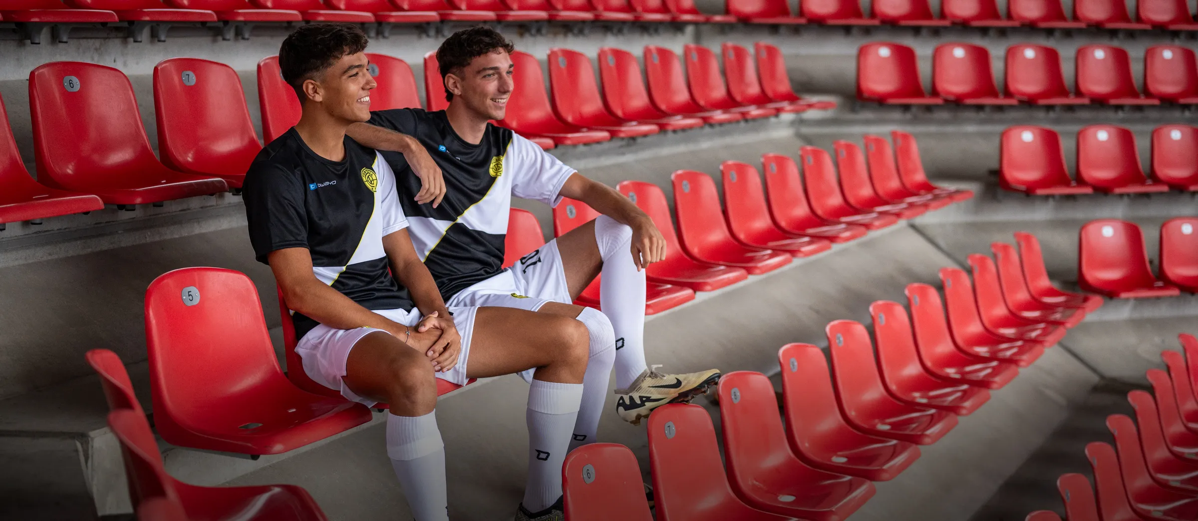 Zwei junge Männer in sportlichen Fußballtrikots sitzen entspannt auf roten Stadionstühlen.