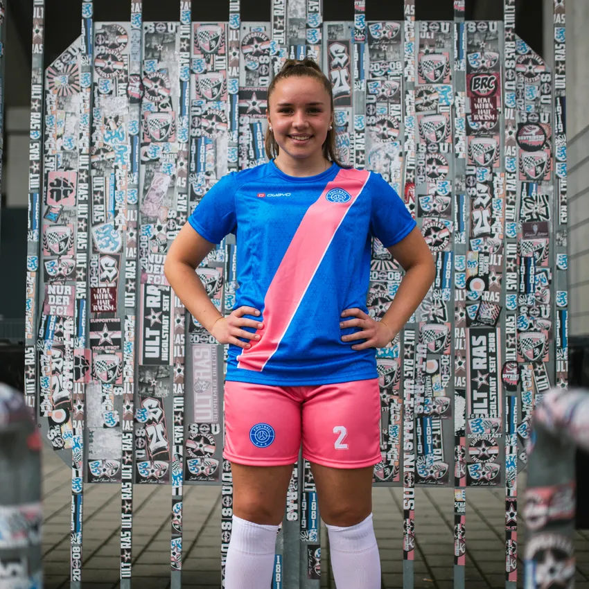 Ein junges Mädchen in einem blauen Fußballtrikot mit pinkem Streifen und pinken Shorts steht vor einer Wand mit Aufklebern.