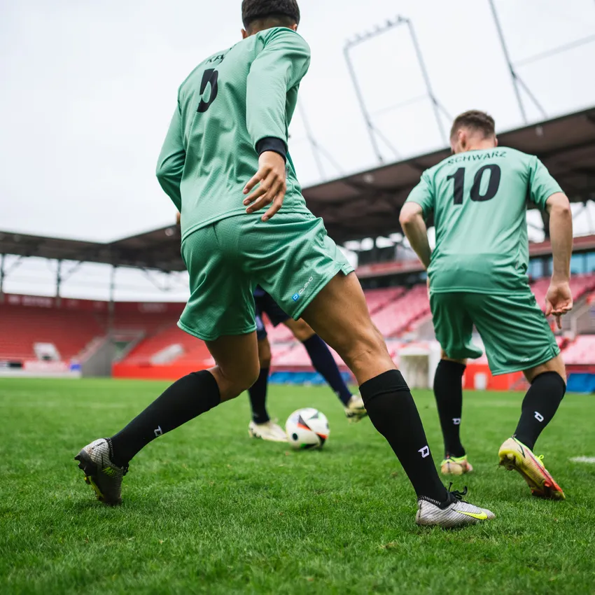 Zwei Fußballspieler in grünen Trikots und schwarzen Socken, die auf einem Rasenfeld mit einem Ball spielen.