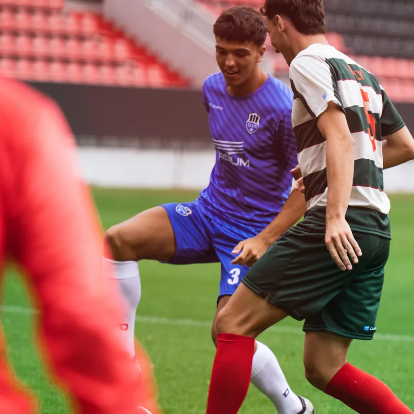 Zwei Fußballspieler in Aktion, einer trägt ein blaues Trikot und der andere ein gestreiftes Trikot in Grün und Weiß, während sie auf einem Spielfeld stehen.