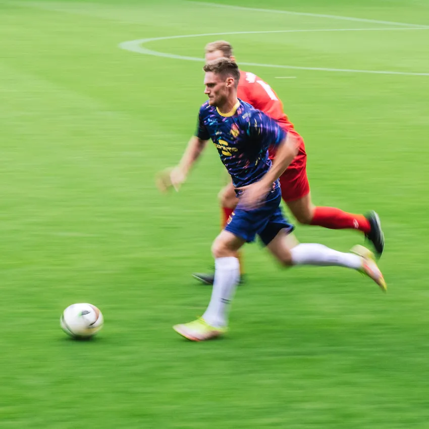 Ein Fußballspieler in einem bunten Trikot sprintet über das grüne Spielfeld, während ein anderer Spieler ihn verfolgt.