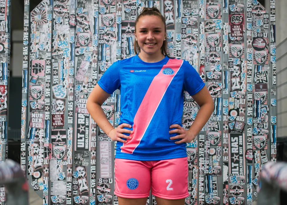 Ein Mädchen steht mit den Händen in die Hüften gestützt in einem blauen Fußballtrikot mit pinkem diagonalen Streifen und pinken Shorts vor einer Wand mit verschiedenen Fußballaufklebern.