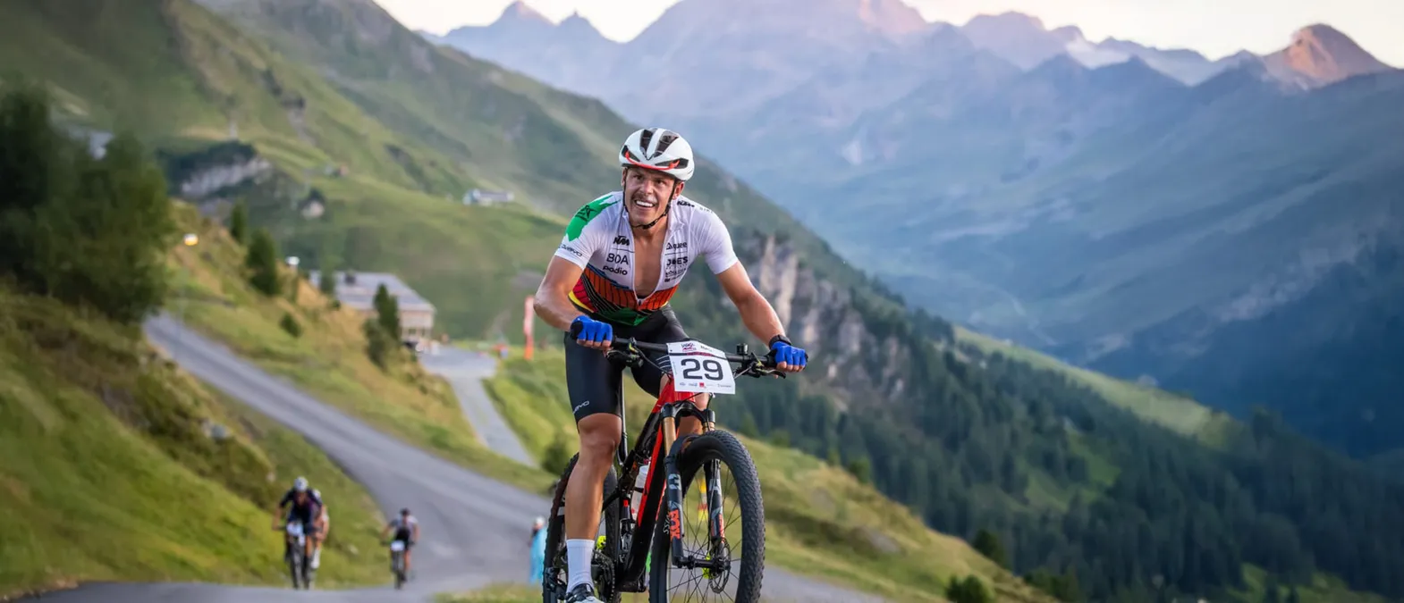 Ein Radfahrer in einem weißen, grünen und orangefarbenen Trikot mit der Nummer 29 fährt auf einem Bergweg in einer bergigen Landschaft.