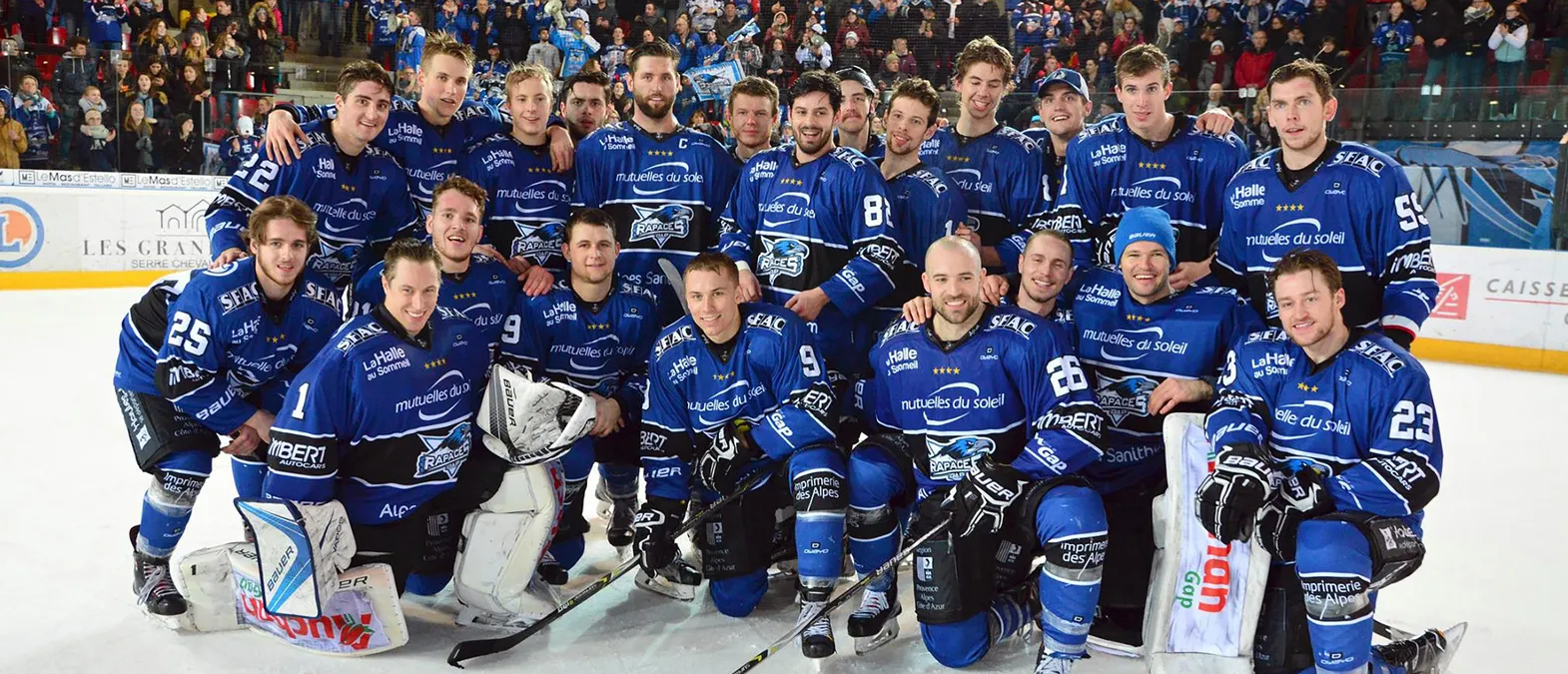 Ein Eishockeyteam in blauen Trikots mit dem Logo der Rapaces de Gap posiert auf dem Eis vor einem Publikum.