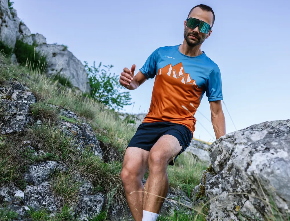 Ein Mann läuft über einen felsigen Weg in einem sportlichen, blau-orangefarbenen T-Shirt und Shorts.