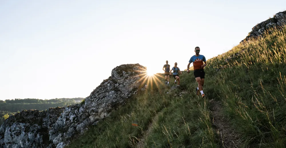 Drei Läufer in funktionaler Sportbekleidung rennen einen hügeligen Weg entlang, während die Sonne hinter ihnen aufgeht.
