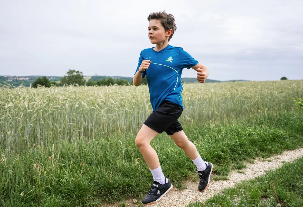 Ein Junge in einem blauen Sportshirt und schwarzen Shorts läuft auf einem Weg durch ein Feld.