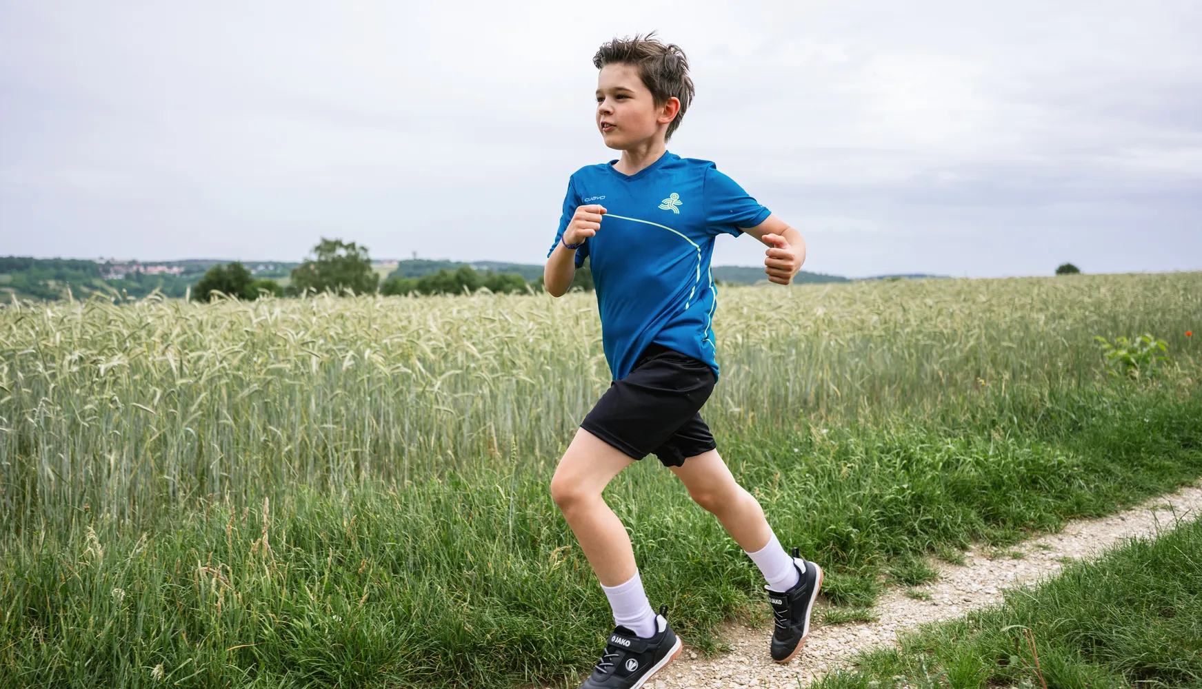 Ein Junge läuft auf einem Weg durch ein Feld, gekleidet in ein blaues Sporttrikot und schwarze Shorts.