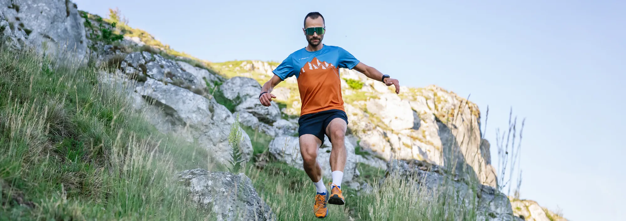 Ein männlicher Läufer in einem orange-blauen Trikot und Shorts, der über einen steinigen Hang läuft.