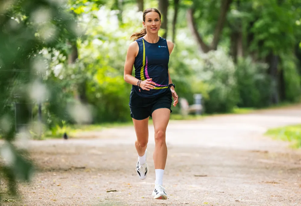 Eine sportliche Frau läuft auf einem Weg im Freien, gekleidet in einem blauen Sporttrikot mit bunten Akzenten und Shorts.