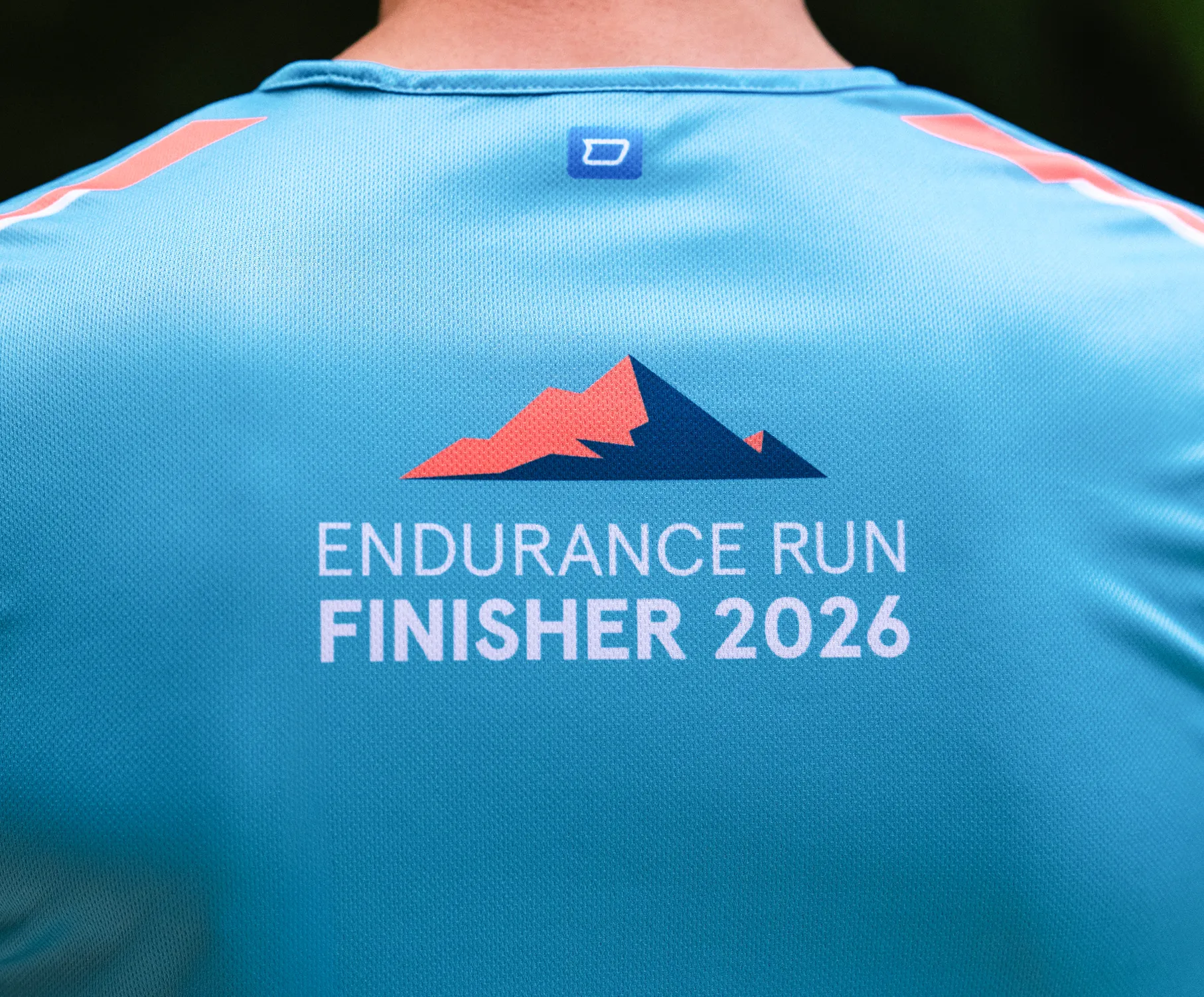 Ein Sportshirt in Blau mit dem Aufdruck 'ENDURANCE RUN FINISHER 2026' und einem stilisierten Bergmotiv auf der Rückseite.