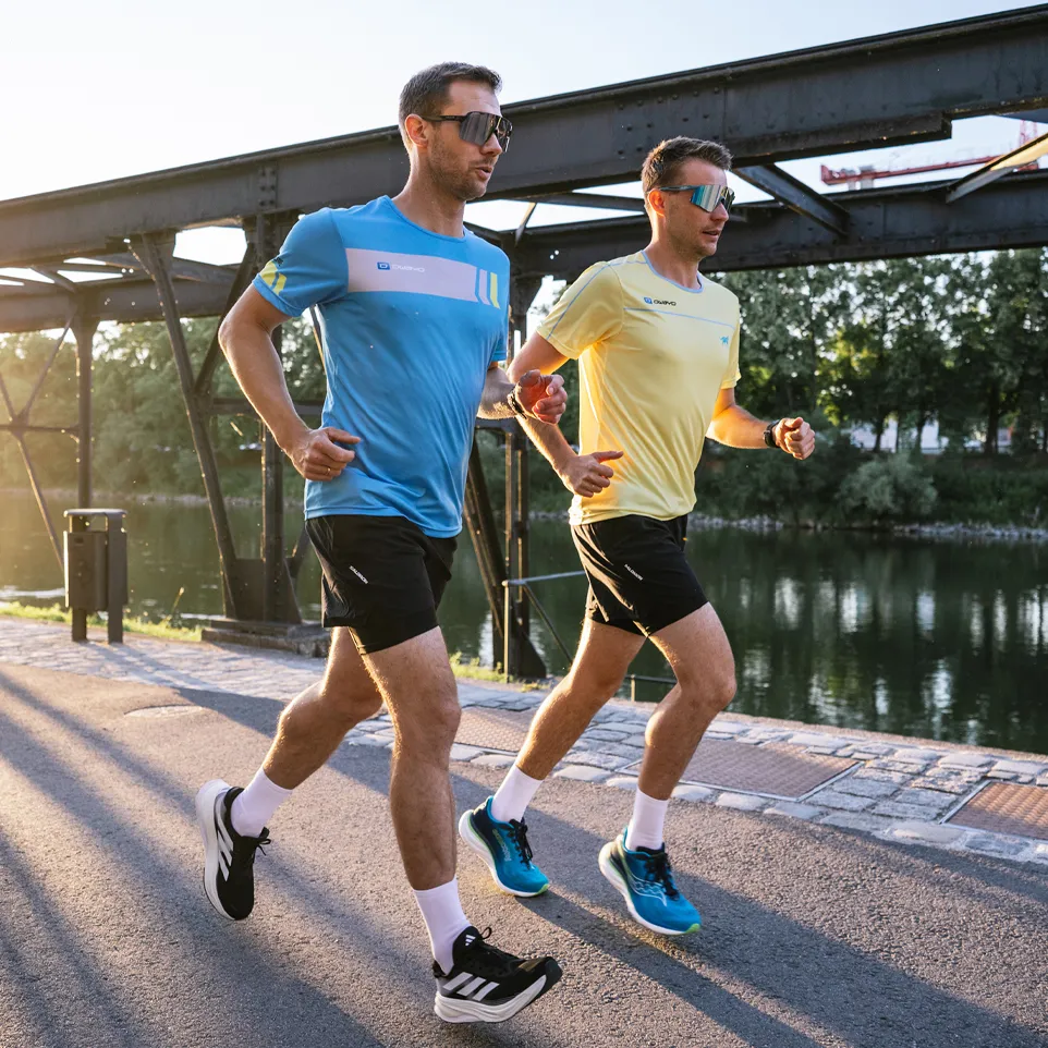 Zwei Männer joggen auf einem Weg entlang eines Gewässers, einer trägt ein blaues T-Shirt und der andere ein gelbes T-Shirt.