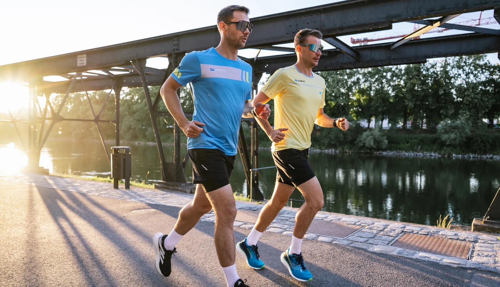 Zwei Männer in sportlicher Laufkleidung, einer in einem blauen T-Shirt und der andere in einem gelben T-Shirt, joggen am Ufer eines Gewässers bei Sonnenuntergang.