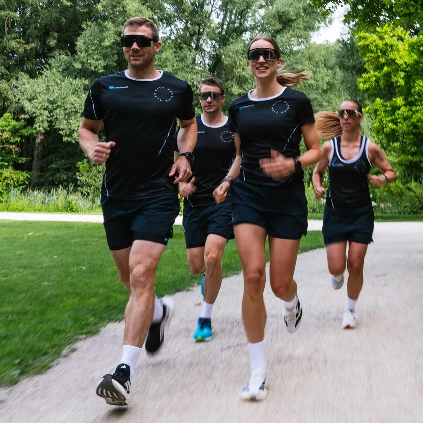 Vier Personen in sportlicher Laufkleidung joggen auf einem Weg durch eine grüne Parklandschaft.