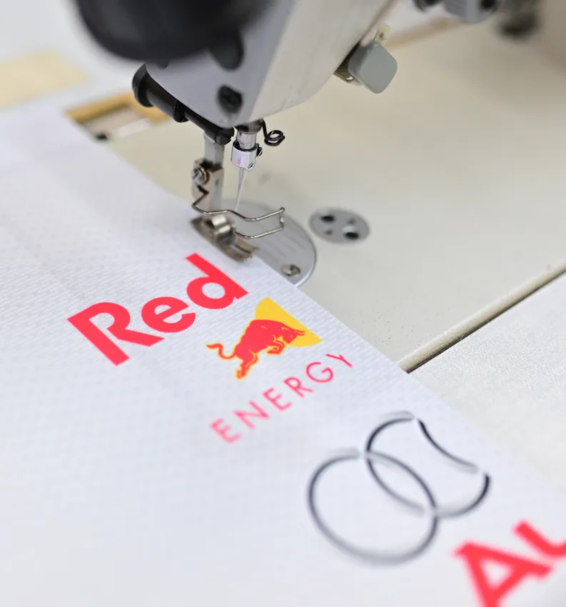 Eine Nähmaschine näht ein weißes Sportbekleidungsstück mit dem roten Red Bull-Logo.
