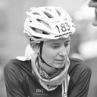 Eine Radfahrerin in einem schwarzen Trikot mit dem Logo 'IWC Schaffhausen', einem weißen Helm mit der Startnummer 183 und einem Schal um den Hals.