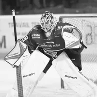 Ein Eishockeyspieler in voller Ausrüstung, einschließlich Helm, Schutzbrille, Handschuhen und Beinschützern, steht in einer Verteidigungsposition auf dem Eis.