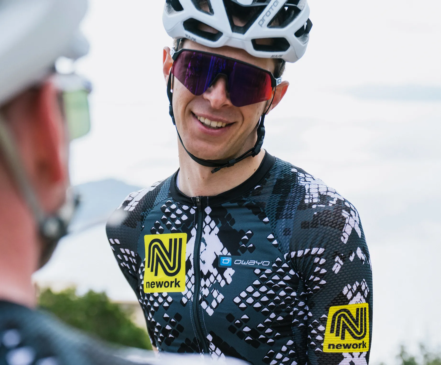 Ein Radfahrer trägt ein maßgeschneidertes, schwarz-weiß gemustertes Trikot mit einem gelben 'nework'-Logo und einem 'owayo'-Logo auf der Brust.