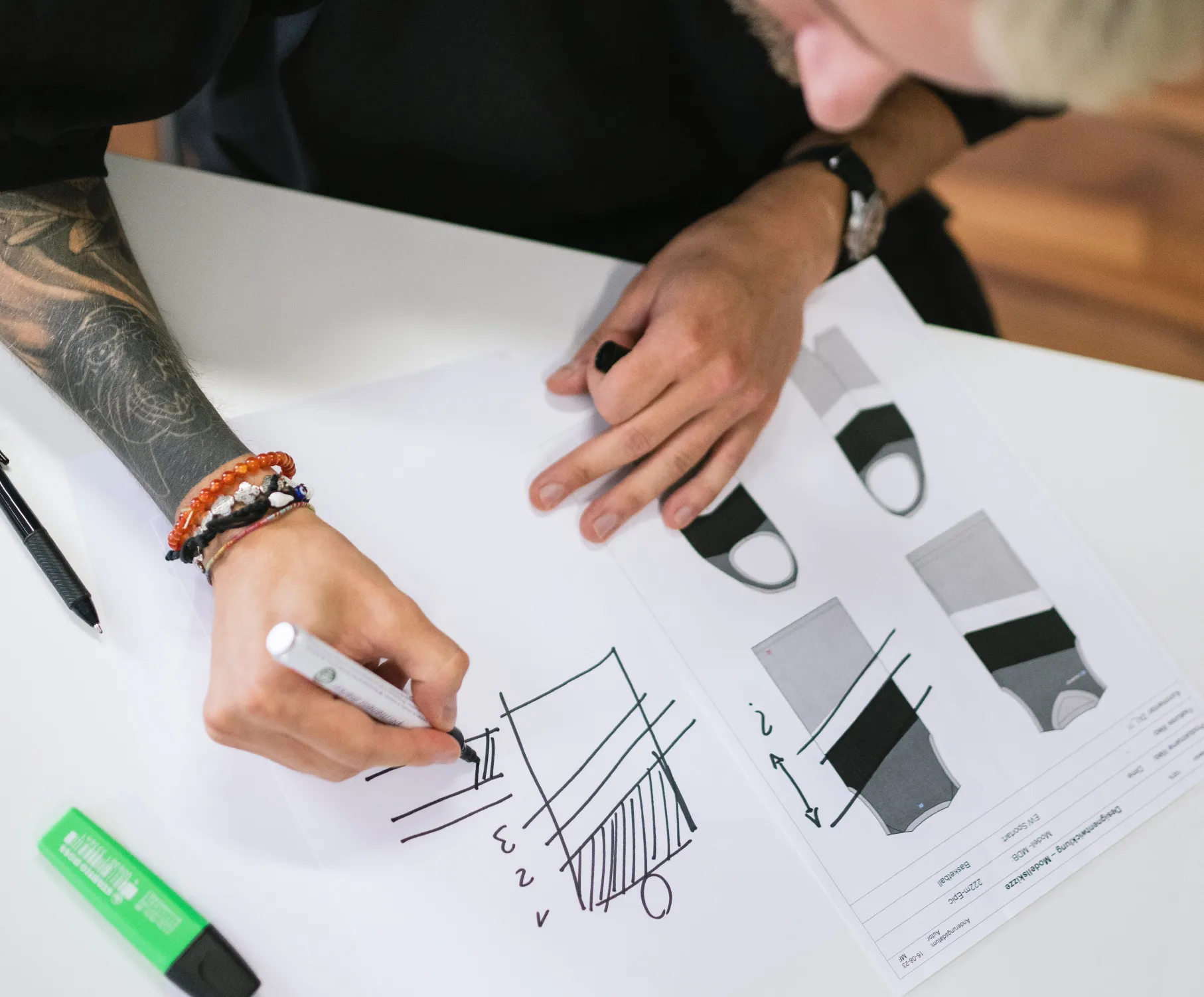 Eine Person mit einem Tattoo am Arm zeichnet mit einem Marker ein Design auf ein Blatt Papier, während auf dem Tisch technische Zeichnungen von Sportbekleidung liegen.
