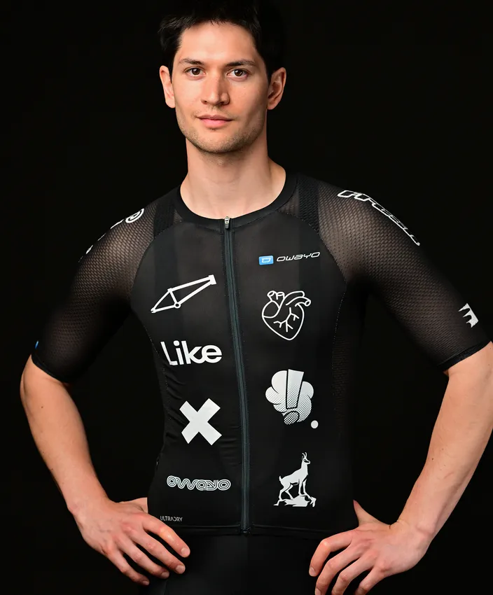 Ein junger Mann trägt ein eng anliegendes, schwarzes Sportoberteil mit verschiedenen weißen Logos und Mustern, posierend vor einem schwarzen Hintergrund.
