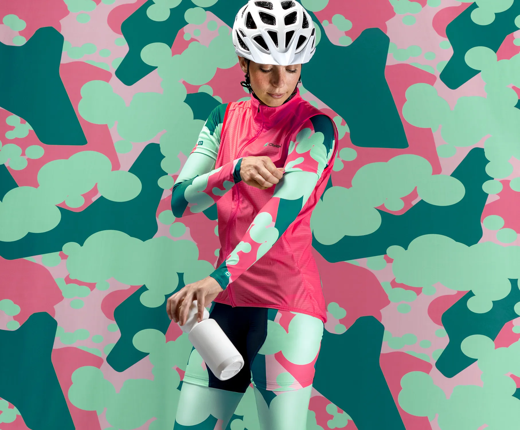 Eine Radfahrerin in einer maßgeschneiderten, pink-grünen Sportbekleidung mit einem abstrakten Muster, die eine weiße Flasche hält und ihre Ärmel anpasst, vor einem farbenfrohen Hintergrund mit dem gleichen Muster.