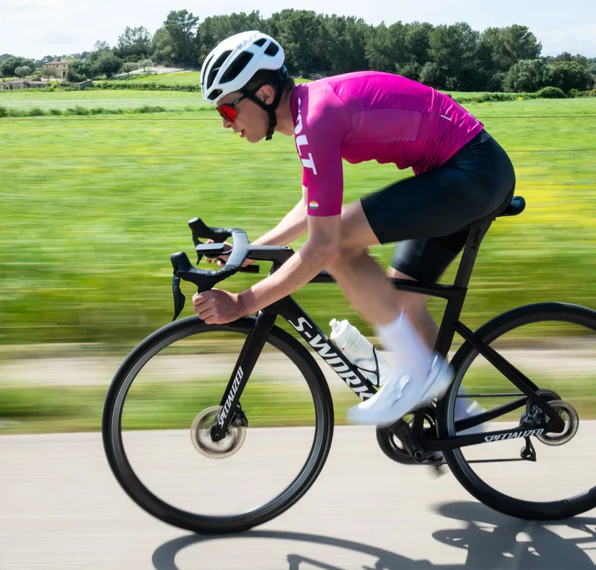 Ein Radfahrer in einem pinken Trikot und schwarzen Shorts fährt auf einer Straße durch eine grüne Landschaft, wobei die Bewegungsunschärfe die Geschwindigkeit betont.